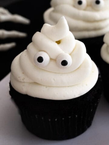 Close up of a black cocoa cupcake with white vanilla buttercream frosting piped to look like a ghost with two candy eyes against a black backdrop.