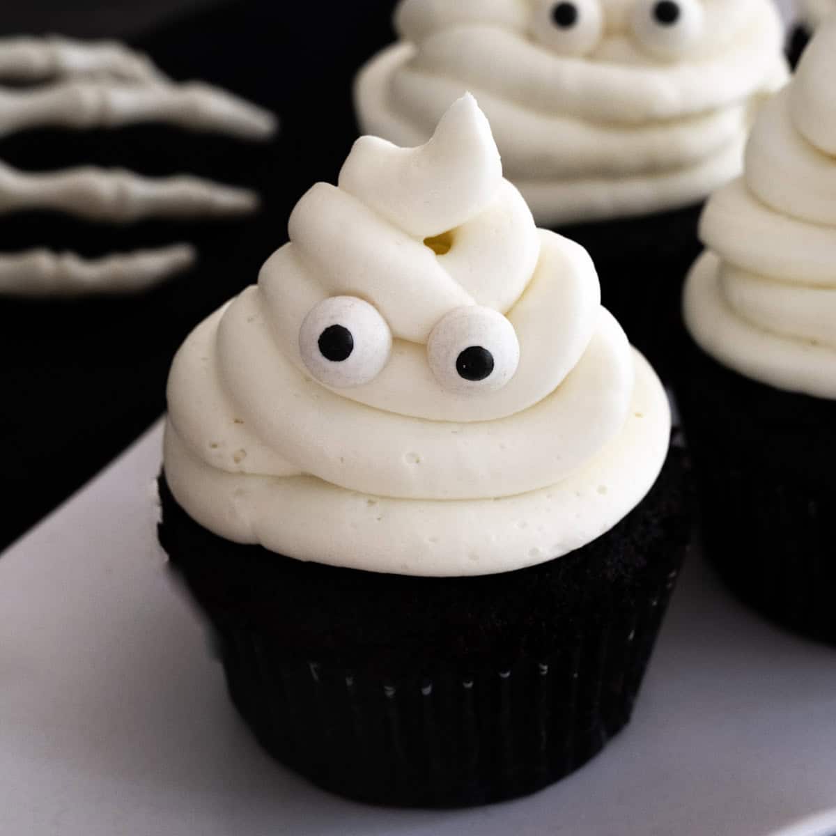 Close up of a black cocoa cupcake with white vanilla buttercream frosting piped to look like a ghost with two candy eyes against a black backdrop.
