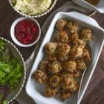 White platter of Thanksgiving turkey meatballs against a dark backdrop with salad, cranberry sauce, mashed potatoes, and gravy around the platter.