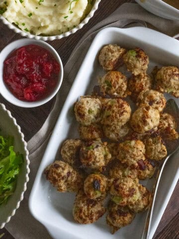 White platter of Thanksgiving turkey meatballs against a dark backdrop with salad, cranberry sauce, mashed potatoes, and gravy around the platter.