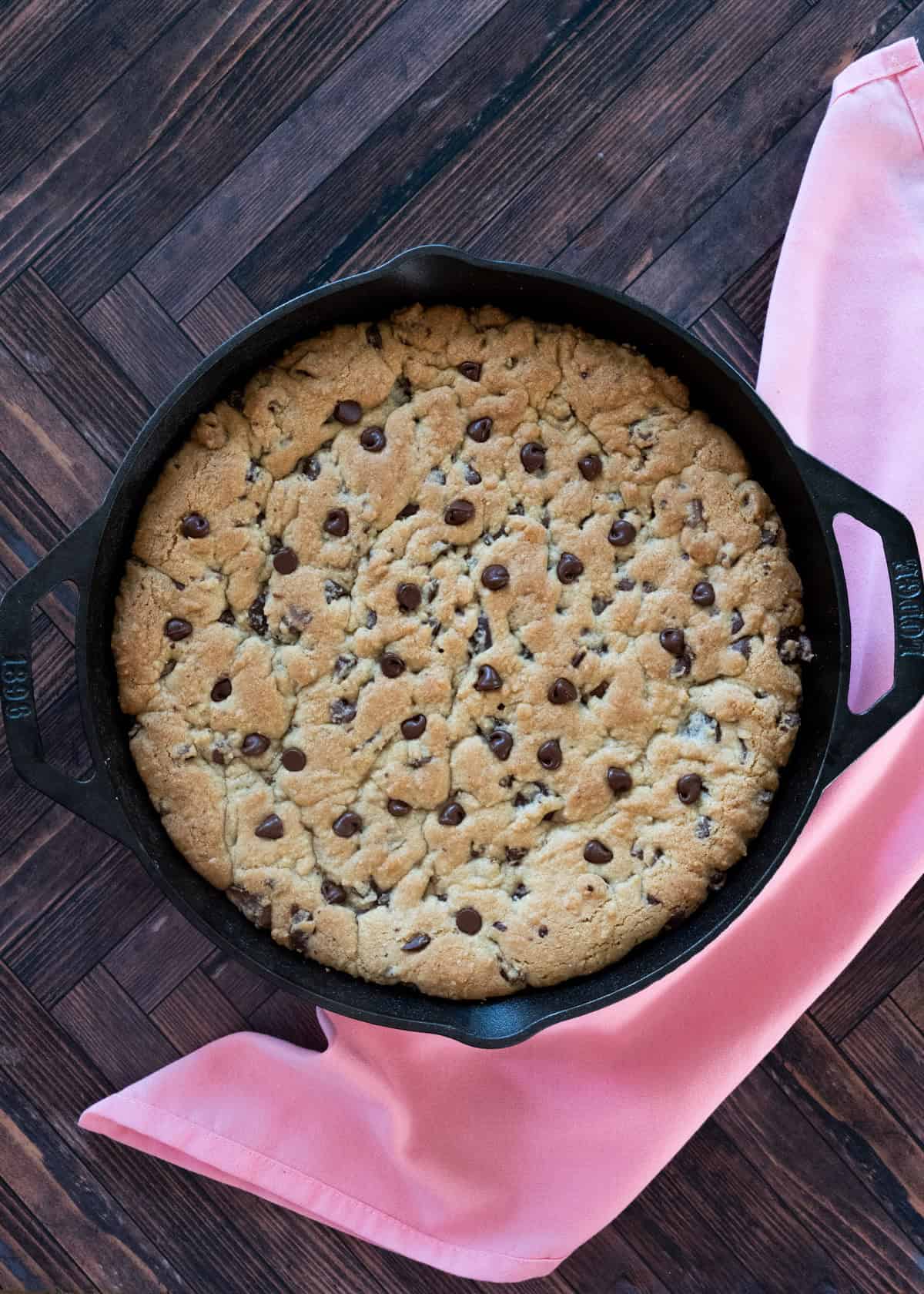 12 inch chocolate chip skillet cookie on a wooden board with a pink napkin surrounding pan.