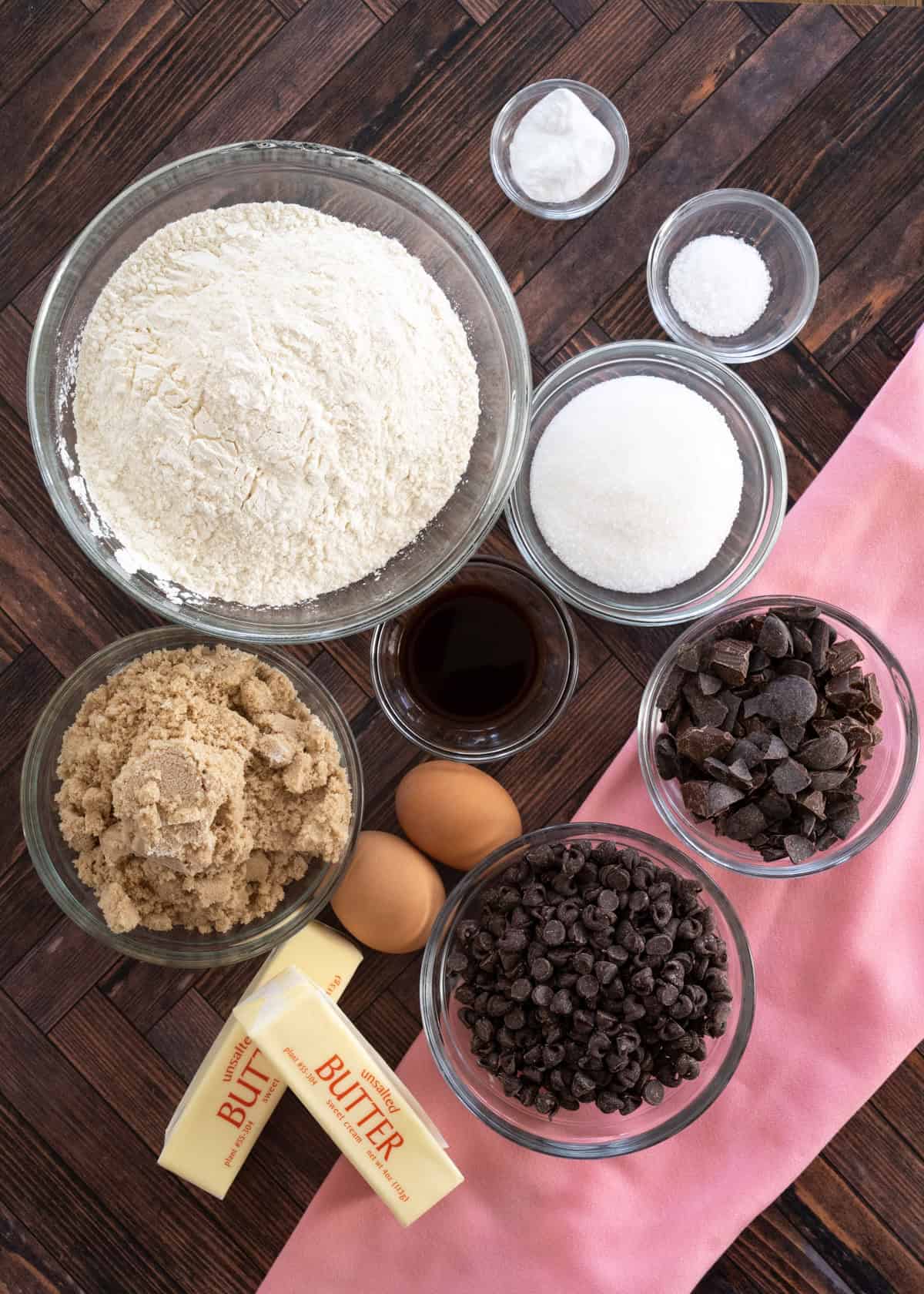 All of the ingredients needed to make a chocolate chp skillet cookie in separate bowls, including flour, granulated sugar, brown sugar, vanilla extract, eggs, butter, baking soda, salt, chocolate chips, and cut up chocolate bar.