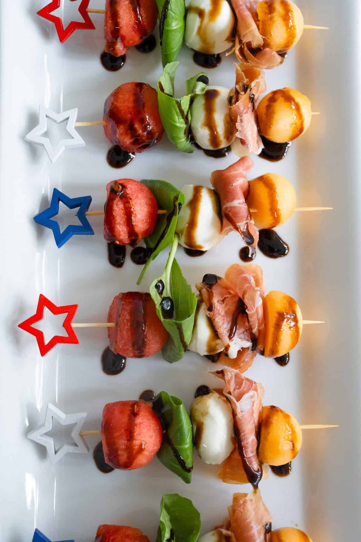 Melon and prosciutto skewers neatly lined up on a white try drizzled with balsamic glaze.