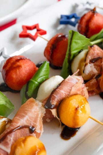 close up of skewers with watermelon, prosciutto, basil, mozzarella, and cataloupe drizzled with balsamic glaze.