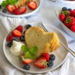 Slice of chiffon cake on a white plate next to a dollop of whipped cream and fresh berries.