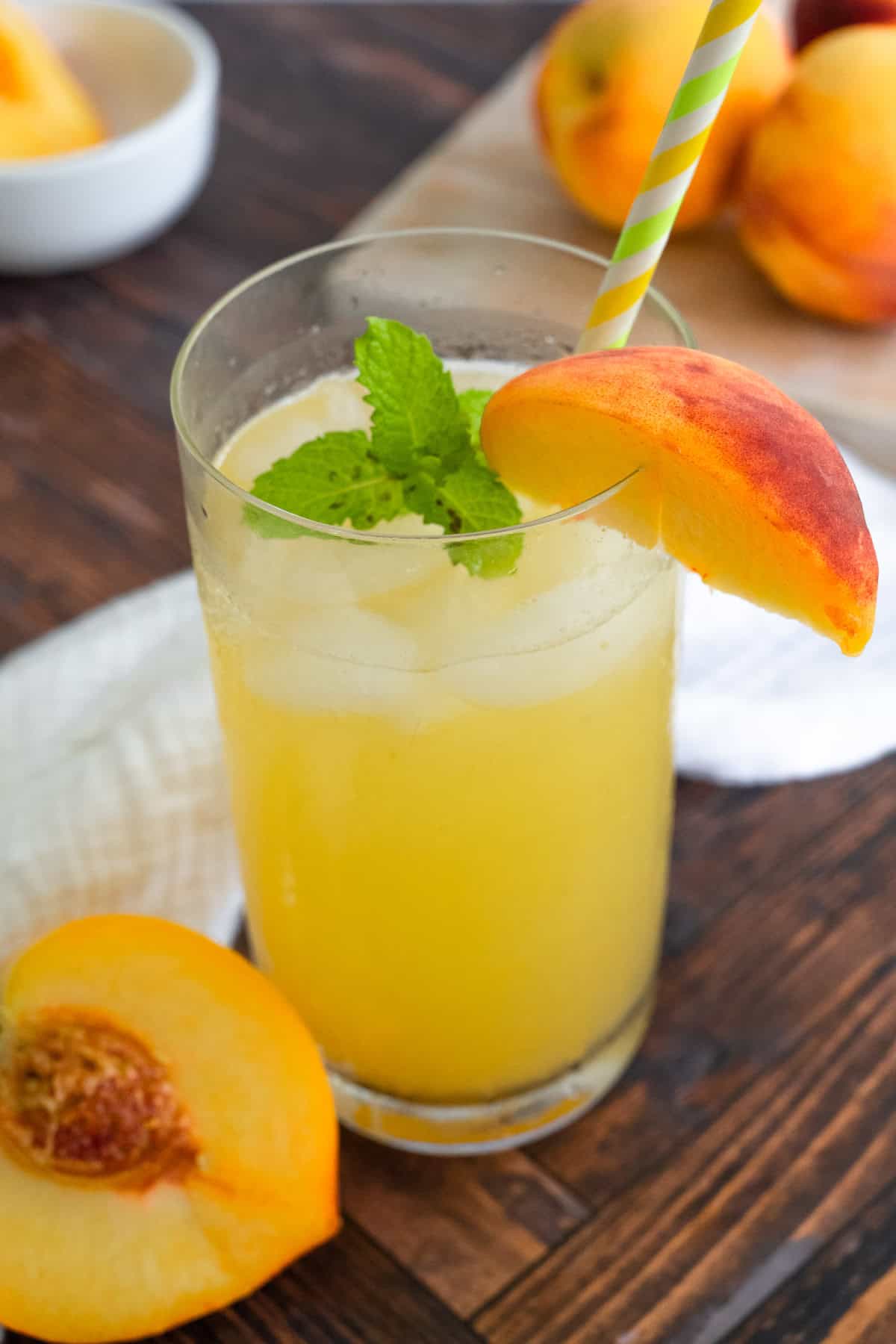 Tall glass of peach lemonade garnished with a peach slice, mint, and a green and yellow-striped straw.